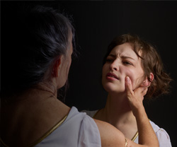 Photo over the shoulder of Jocasta holding Antigone's cheek as Antigone recoils from her