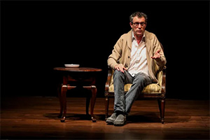 Frank Blocker seated in an armchair on a dark stage to address the audience in GOOD JEW